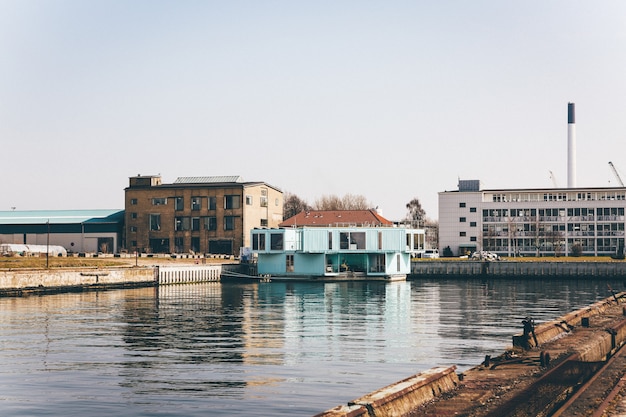 Wide shot of a light blue house on a dock on the body of water near buildings under a clear sky