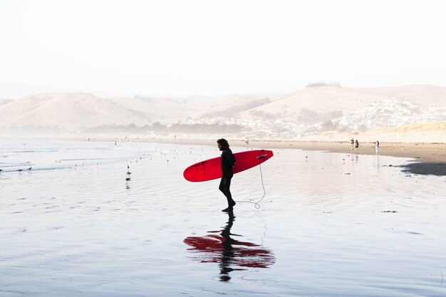 Широкий выстрел мужской серфер в костюме для серфинга с доской для серфинга на берегу моря