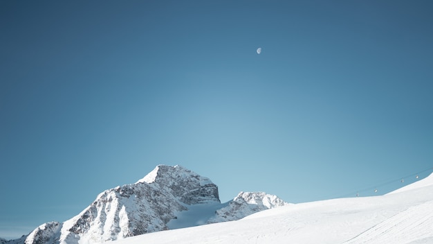 Wide shot of a mountain covered in snow under a clear blue sky with a half moon
