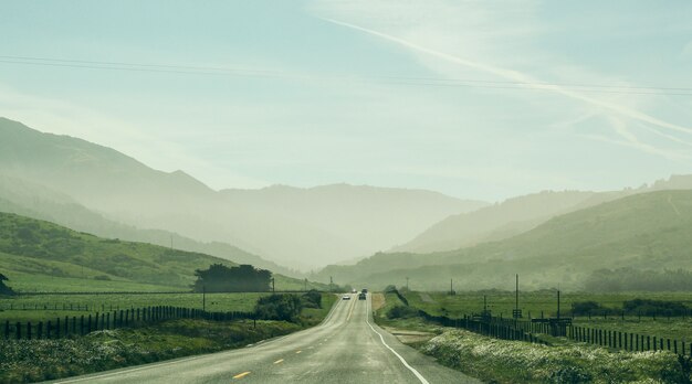 Wide shot of a road in the middle of a grassy field with cars driving and a forested mountain