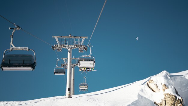 Wide shot of ropeways and gray pillar on a snowy surface under a clear blue sky with a half moon