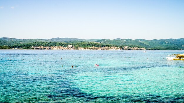 Wide shot of a sea with buildings on the shore and forested mountains in the distance