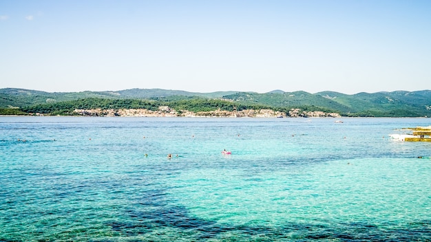 Wide shot of a sea with buildings on the shore and forested mountains in the distance