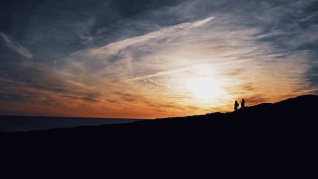 Wide shot of silhouettes of two people walking on a hill with the sun shining