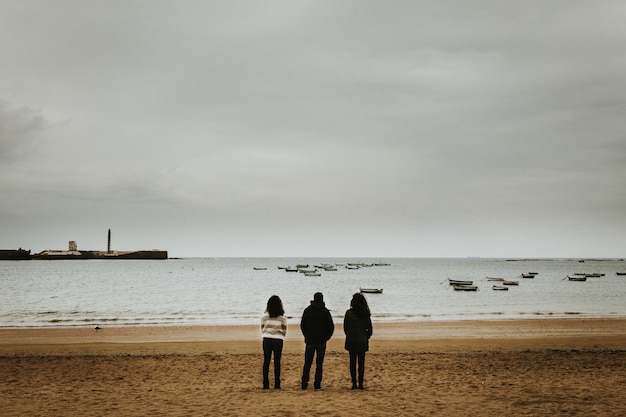 Wide shot of three people standing near the seashore with small boats floating in the sea