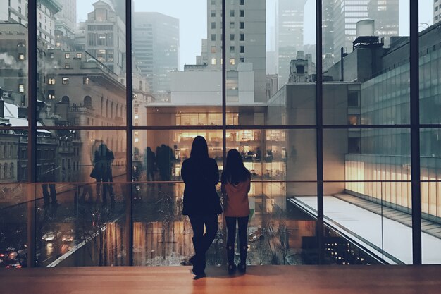 Wide shot of two females standing at a huge glass window looking at the view of high rise buildings