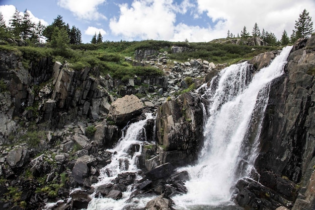 Wide shot of a waterfall from a cliff with grass and trees on the surface on a cloudy day