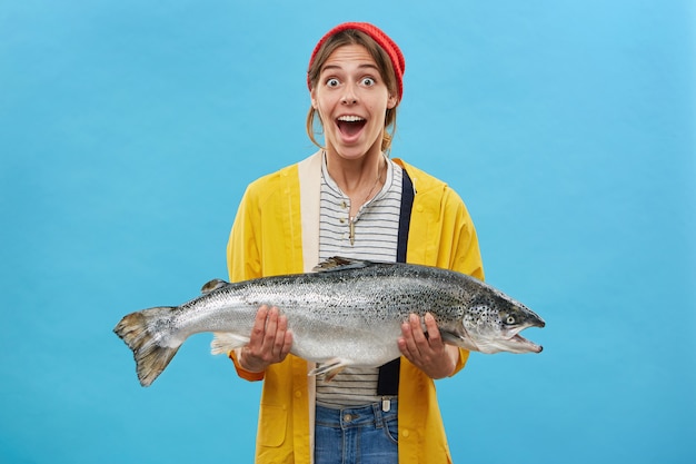 Free photo wife of fisherman holding huge fish having surprised expression looking with bugged eyes and jaw dropped not believing her eyes rejoicing successful catch. happy shocked fisherwoman with trout