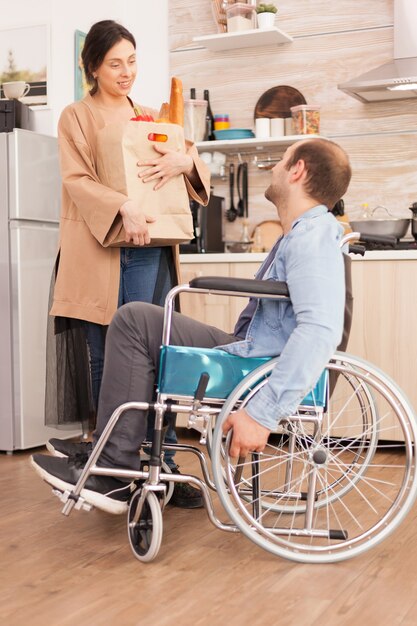 Wife holding paper bag with organic products in kitchen talking with disabled husband in wheelchair. Disabled paralyzed handicapped man with walking disability integrating after an accident.