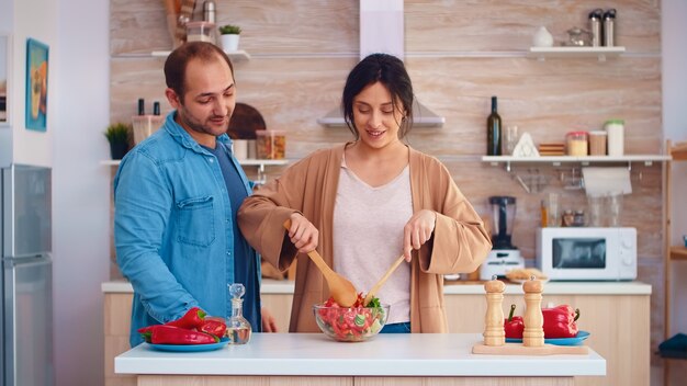 Wife mixing healthy salad on glass bowl and husband with grocery paperbag in kitchen. Cooking preparing healthy organic food happy together lifestyle. Cheerful meal in family with vegetables