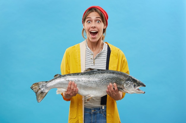 Бесплатное фото Жена рыбака держит в руках огромную рыбу с удивленным выражением лица, с вытаращенными глазами и отвисшей челюстью, не веря своим глазам, радуясь удачному улову. счастливый шокирован рыбак с форелью