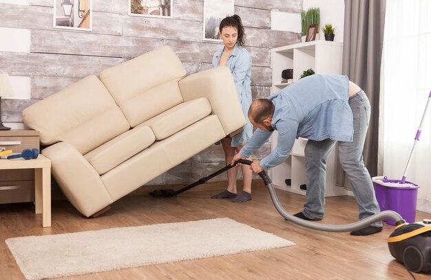 Жена поднимает диван, пока муж чистит пыль под ним пылесосом