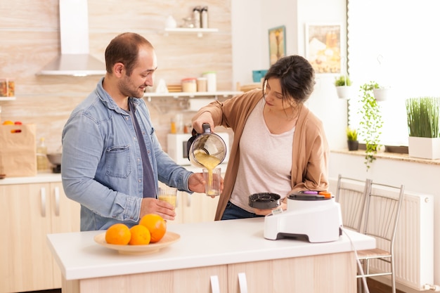 Wife pouring tasty smoothie while husband is holding the glass. Healthy carefree and cheerful lifestyle, eating diet and preparing breakfast in cozy sunny morning