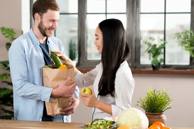 Wife taking vegetable from husband holding brown grocery bag in kitchen
