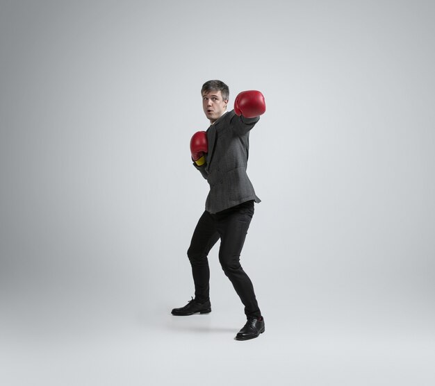 Wild and young. Caucasian man in office clothes boxing with two red gloves on grey wall. Businessman training in motion, action. Unusual look for sportsman, activity. Sport, healthy lifestyle.