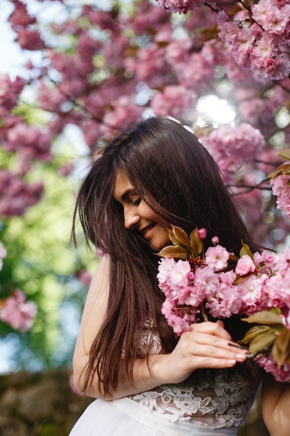 Ветер дует волосы брюнетки женщины, пока она ставит перед цветущим деревом сакуры