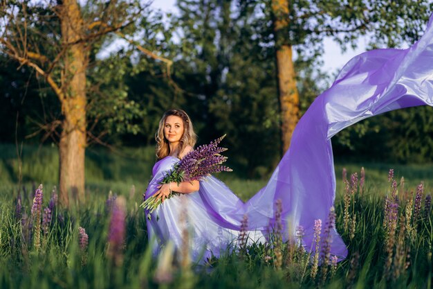 Wind blows pregnant woman's violet dress while she stands in the field of lavender