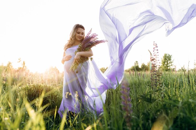 彼女がラベンダーの畑に立っている間、風は妊婦の紫色のドレスを吹き飛ばします