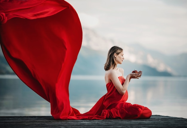 Wind blows red dress of a pregnant woman sitting with apple on the bridge over the lake