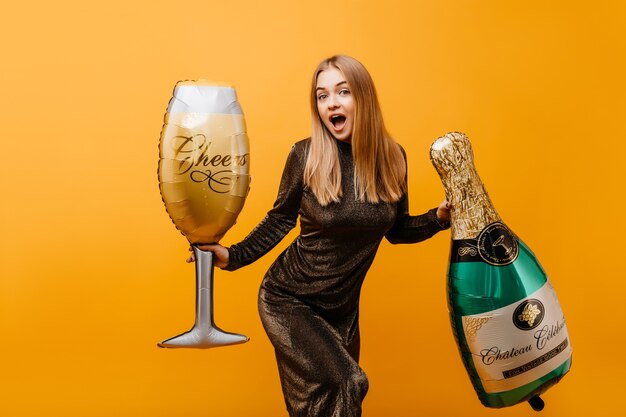 Winderful woman with straight hairstyle expressing surprised emotiins at birthday party. Indoor portrait of beautiful graceful woman with bottle of champagne and wineglass.