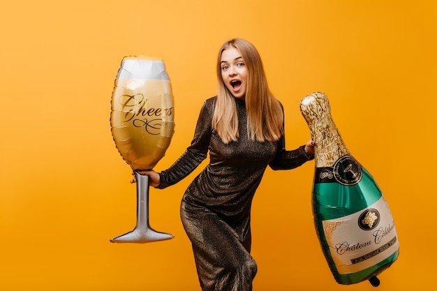 Winderful woman with straight hairstyle expressing surprised emotiins at birthday party. Indoor portrait of beautiful graceful woman with bottle of champagne and wineglass.