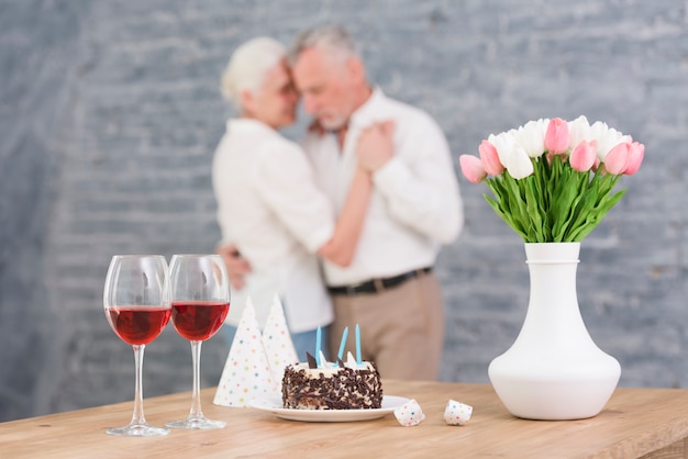 Foto gratuita bicchiere di vino; cappello da festa; torta di compleanno e vaso di fiori sul tavolo davanti ballare coppia offuscata