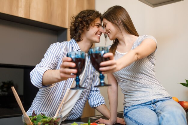 Время вина, молодая супружеская пара улыбается, пьет красное вино и готовит вместе на кухне