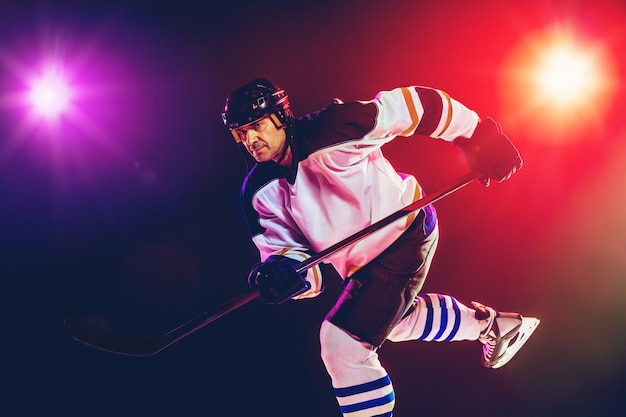 Winner. Male hockey player with the stick on ice court and dark neon colored wall. Sportsman wearing equipment, helmet practicing. Concept of sport, healthy lifestyle, motion, wellness, action.