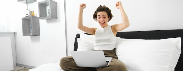 Foto gratuita ragazza vincente che guarda il portatile con le mani alzate seduta sul letto guardando qualcosa nel portatile e