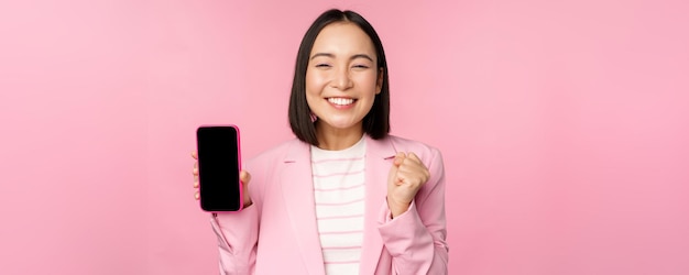 Winning korean businesswoman showing smartphone screen smiling pleased demonstrating mobile phone application online store or shopping app standing over pink background