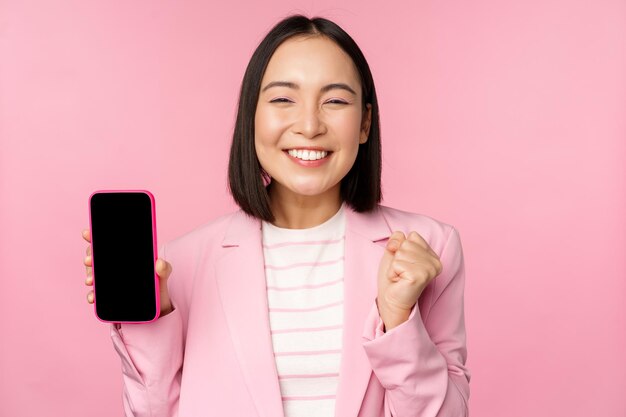 스마트폰 화면을 보여주는 수상한 한국 여성 사업가가 핑크색 배경 위에 서 있는 휴대폰 앱 온라인 스토어 또는 쇼핑 앱을 시연하며 기뻐했습니다.