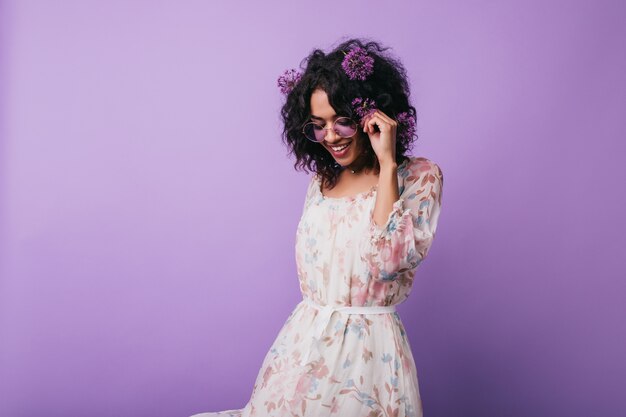 Winsome african girl posing in dress with floral pattern. Portrait of carefree black lady with alliums in hair.