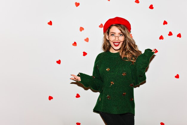 Winsome brown-haired female model in trendy green sweater fooling around on indoor photoshoot