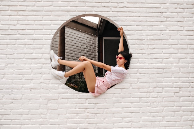 Free photo winsome caucasian woman sitting on bricked wall. outdoor shot of glad tanned woman posing on urban background.