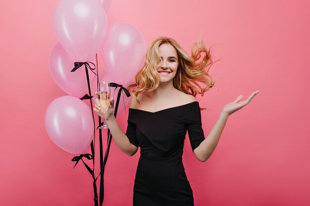 Очаровательная европейская девушка с волнистыми волосами празднует день рождения. Милая молодая женщина с гелиевыми шарами, танцующими на вечеринке.