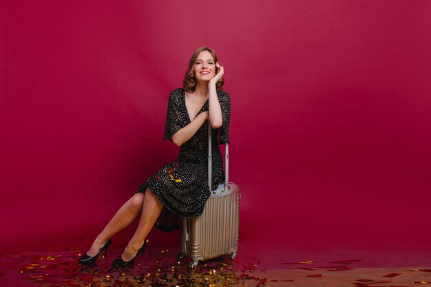 Winsome girl in high heel shoes sitting on suitcase after party with friends