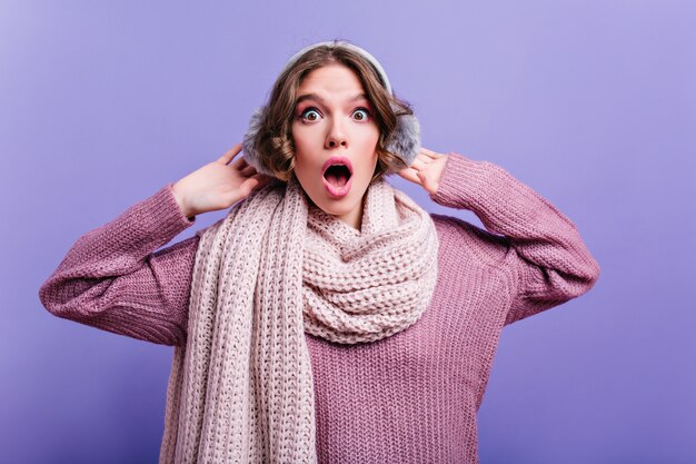 Winsome girl with dark hair wears warm scarf expressing amazement with mouth open. Indoor close-up photo of curly shocked girl in fur headpones.