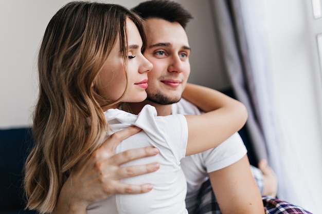 Winsome girl with long hair embracing her boyfriend with eyes closed. Indoor portrait of cute married couple spending weekend together.