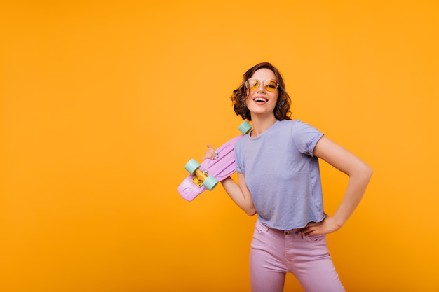 Winsome girl in yellow-colored glasses posing. Dark-haired blithesome lady with skateboard standing.