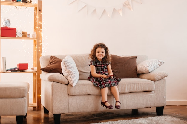 Winsome kid sitting in living room. Indoor shot of preteen girl in dress posing on couch.