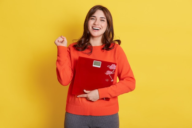 Winsome lady with floor scales in hands posing over yellow, clenching fist, being glad to slim down, finely loosing weight, girl wearing casual attire.