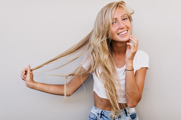 Winsome tanned woman in tank-top touching her long hair. Good-humoured blonde woman posing on white background.