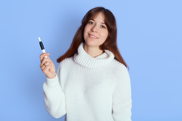 Winsome vaping young woman over blue, lady with good mood looks at camera and holds e ciggy in her hand, wearing casual attire, has dark hair.