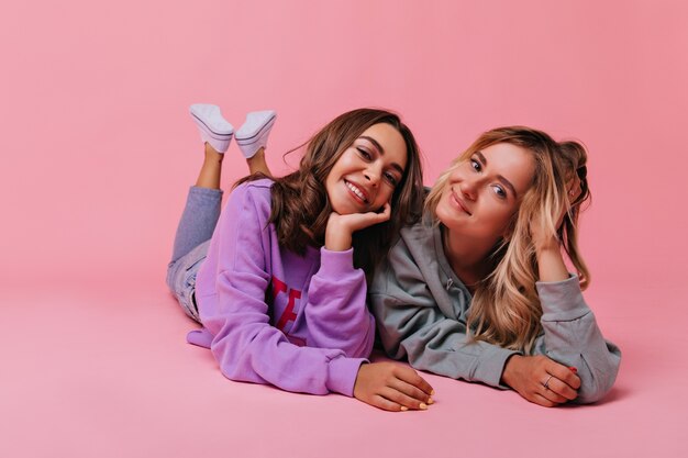 Winsome white ladies lying on the floor with smile. Cute female friends chilling with pink interior.