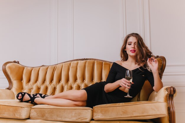 Привлекательная женщина в черном длинном платье, лежа на коричневом диване с серьезным выражением лица. Крытый портрет пугающей девушки с элегантной прической, пьющей вино на софе.