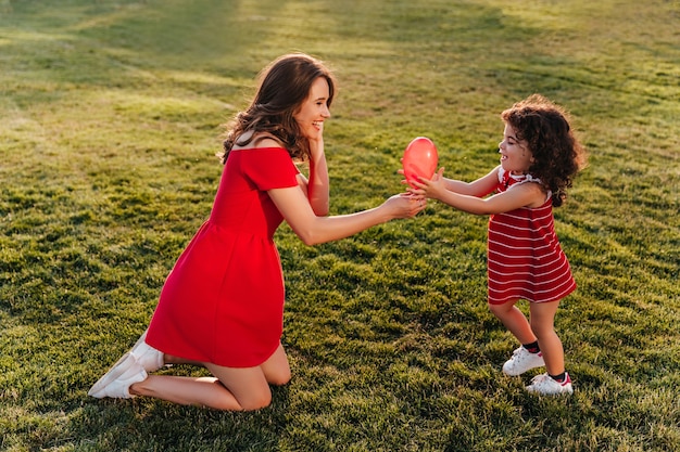 Winsome woman in red dress playing with her daughter in park. Outdoor photo of laughing young lady looking at little sister with smile.