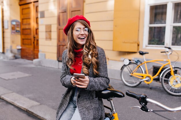 Winsome woman in trendy tweed coat looking away with laugh in front of yellow house