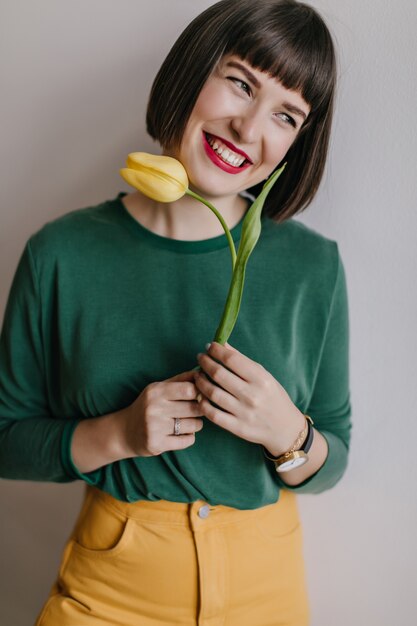 Обаятельная женщина с короткими темными волосами позирует с желтым тюльпаном. Крытый портрет восторженной девушки в зеленой рубашке с цветком и смехом.