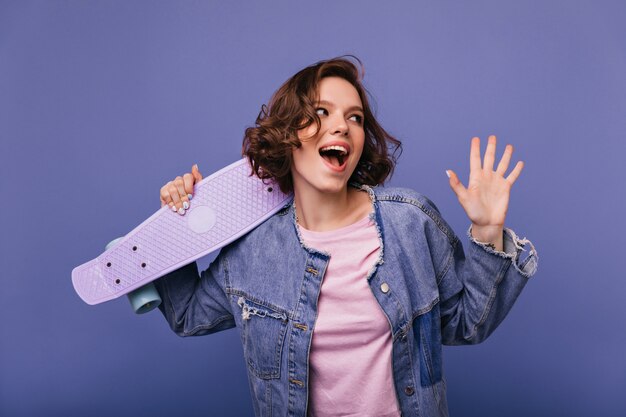 Winsome young woman with pretty smile posing with skateboard. Indoor shot of laughing carefree lady with short wavy hair standing.