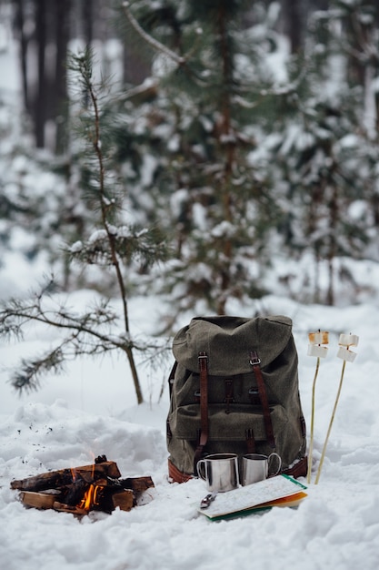冬のキャンプ。旅行のリュックサック、地図、雪の上に2つのマグカップとマシュマロと一緒にボンファイア